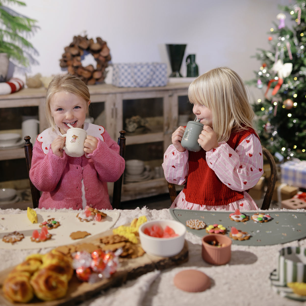 Two girls smile, sipping from Mushie Silicone Training Cups + Straws at a festive table.