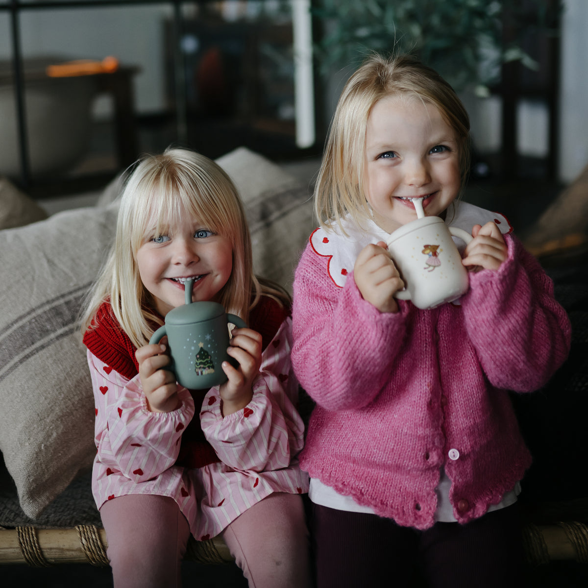 Two girls enjoy drinks from Mushie Silicone Training Cup + Straw while sitting on a couch.