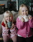 Two girls enjoy drinks from Mushie Silicone Training Cup + Straw while sitting on a couch.