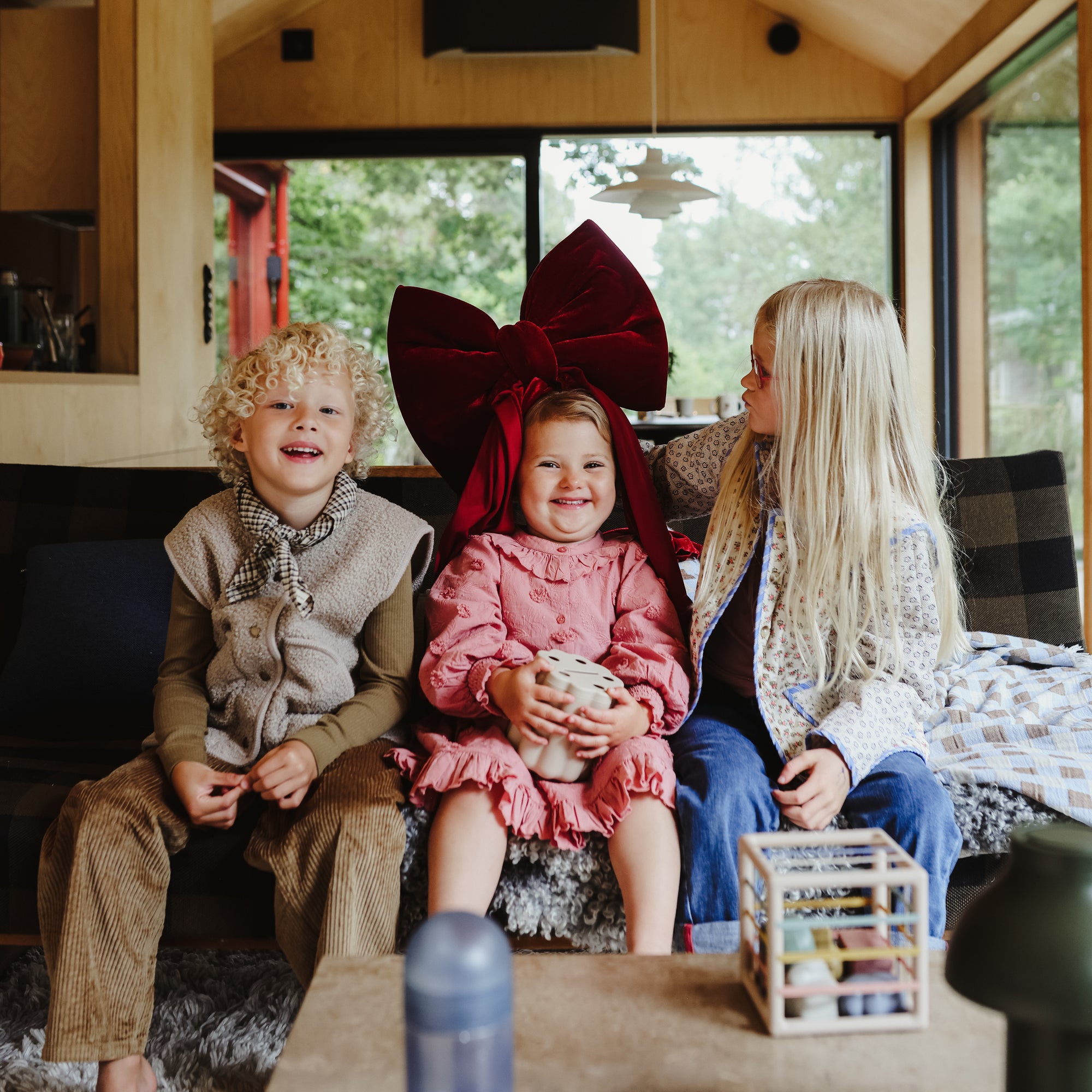 Three young children sit on a couch in a cozy room. The center child wears a large red bow, smiling as the others sit nearby. A mushie Coin &amp; Tube Sorting Set and a water bottle are on the table in front of them.