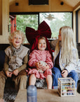 Three young children sit on a couch in a cozy room. The center child wears a large red bow, smiling as the others sit nearby. A mushie Coin & Tube Sorting Set and a water bottle are on the table in front of them.
