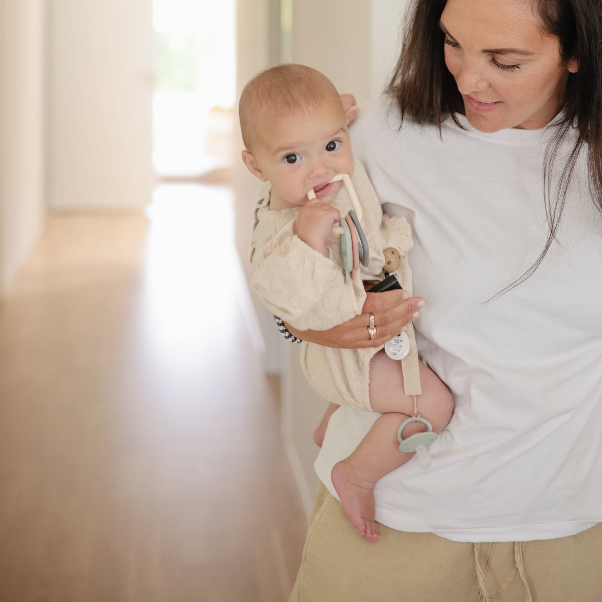 A woman smiles down at her baby, who is dressed in light clothes and chewing on a FRIGG Little Viking Natural Rubber Pacifier by FRIGG, attached to their outfit, while looking at the camera in a hallway.