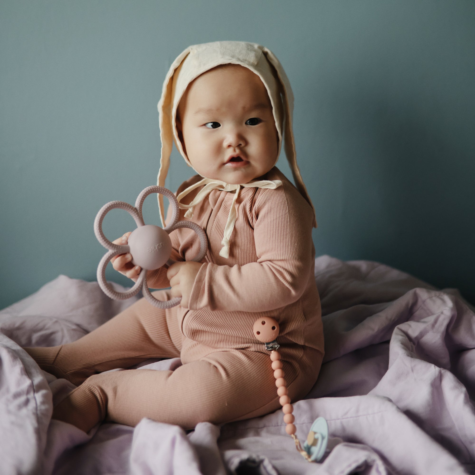 A baby in a pink outfit and bunny ear bonnet sits on a lavender blanket, holding a flower-shaped teether and a FRIGG Little Viking Natural Rubber Pacifier 2-Pack by FRIGG, made in Denmark.