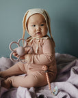 A baby in a pink outfit and bunny ear bonnet sits on a lavender blanket, holding a flower-shaped teether and a FRIGG Little Viking Natural Rubber Pacifier 2-Pack by FRIGG, made in Denmark.