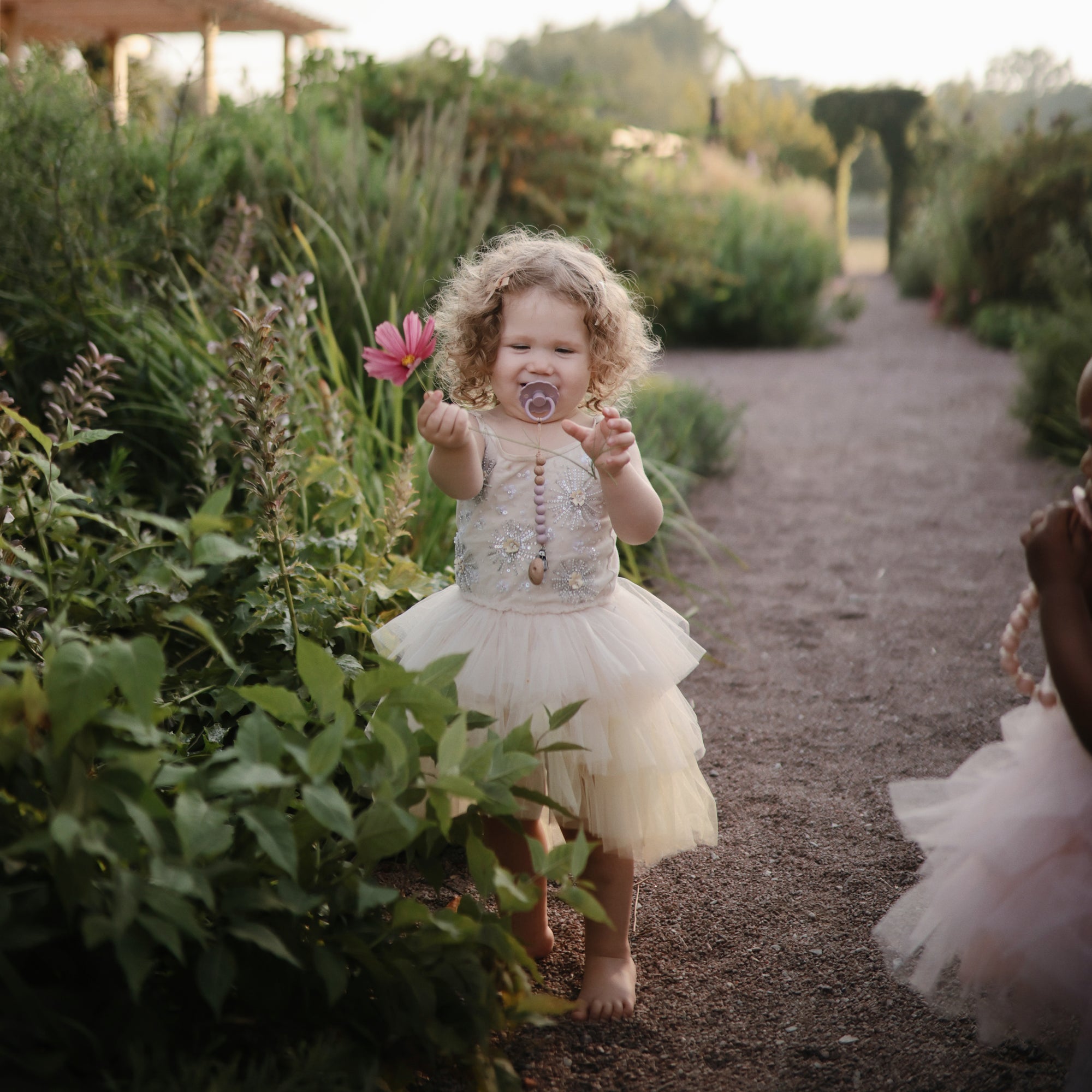 Curly-haired child in a tutu with a FRIGG Daisy Natural Rubber Pacifier and pink flower in the garden.
