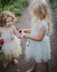 Two girls in frilly dresses stand barefoot, holding flowers and a FRIGG Daisy rubber pacifier.