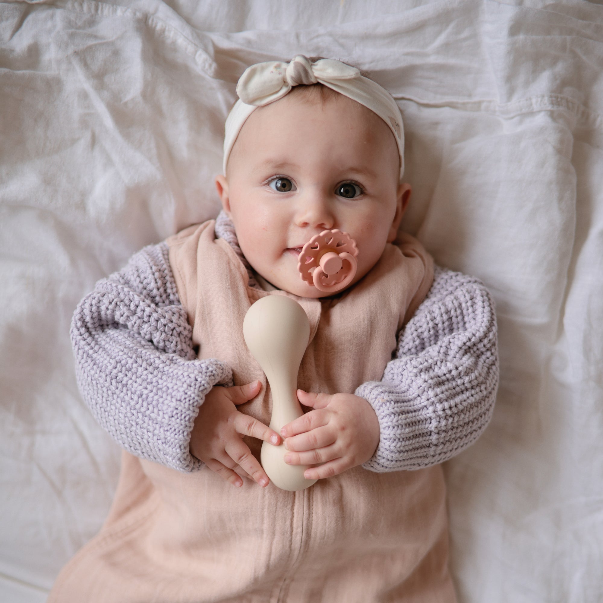 A baby in a light pink outfit and gray knit sweater lies on a white blanket, wearing a headband and holding a beige rattle, using the FRIGG Andersen Fairytale Silicone Pacifier 2-Pack by FRIGG, made safely in Denmark.