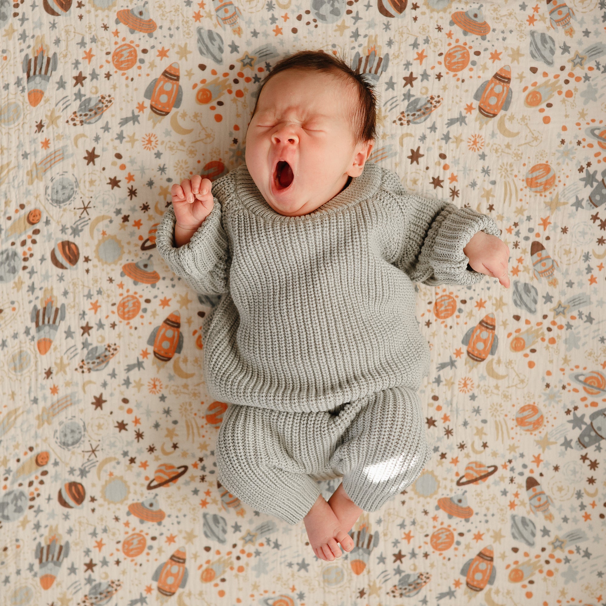 A baby in a cozy gray Mushie Chunky Knit Beanie and an organic cotton outfit lies yawning on a patterned blanket with space-themed designs, eyes closed and one fist slightly raised.