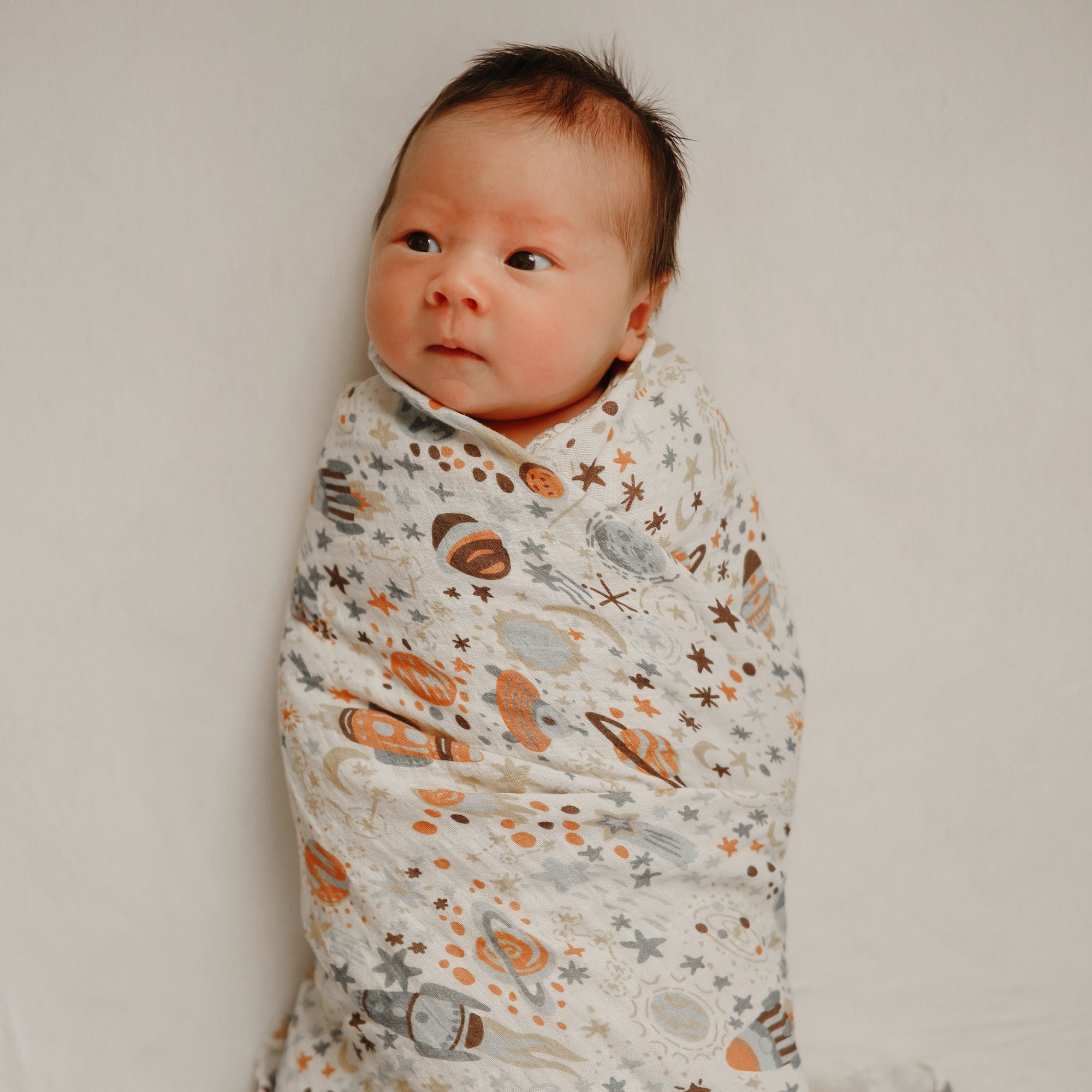 A baby lies on a soft white surface, wrapped in a mushie Organic Cotton Muslin Swaddle Blanket featuring orange, gray, and brown space-themed patterns, gazing slightly to the side.