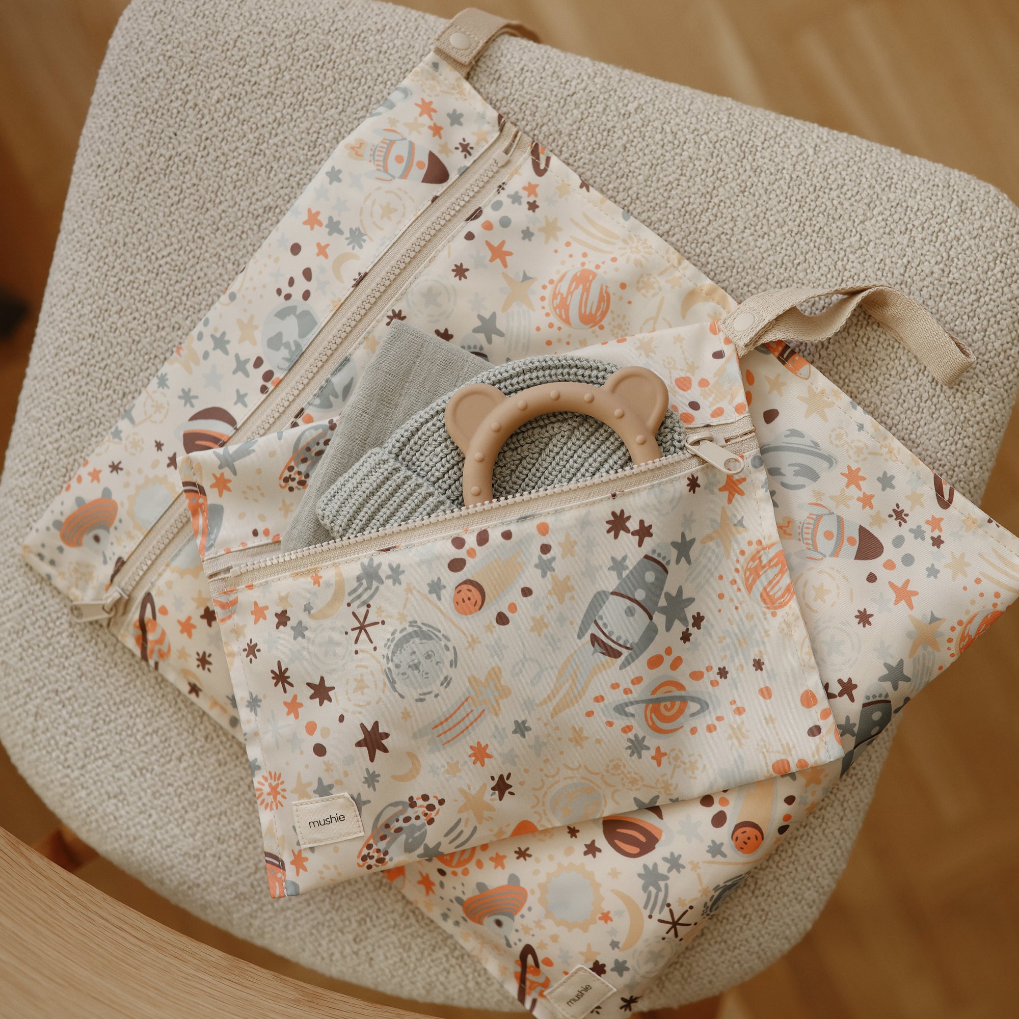 A Mushie Chunky Knit Beanie rests on a beige chair, with a gray cloth and a tan baby teething ring with bear ears partially visible inside the pouch.