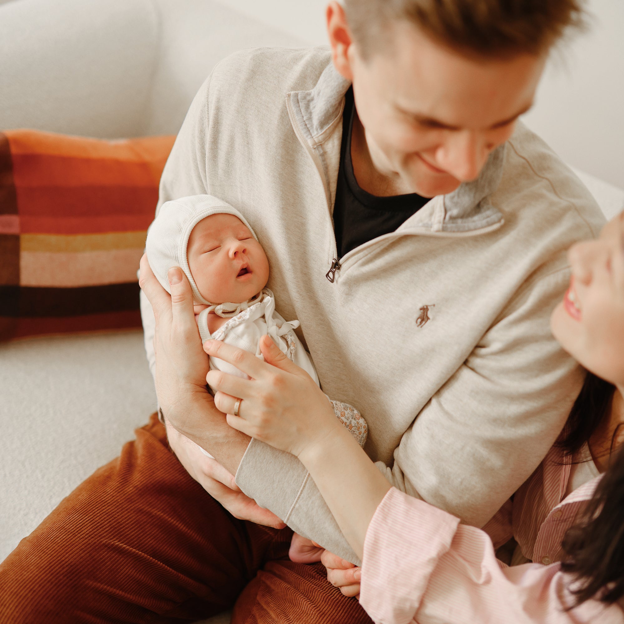 A man cradles a sleeping newborn in his arms, the baby cozy in a mushie Ribbed Baby Bonnet. A woman gently touches his arm as they sit closely together on a white couch with a colorful pillow in the background.