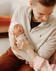 A man cradles a sleeping newborn in his arms, the baby cozy in a mushie Ribbed Baby Bonnet. A woman gently touches his arm as they sit closely together on a white couch with a colorful pillow in the background.