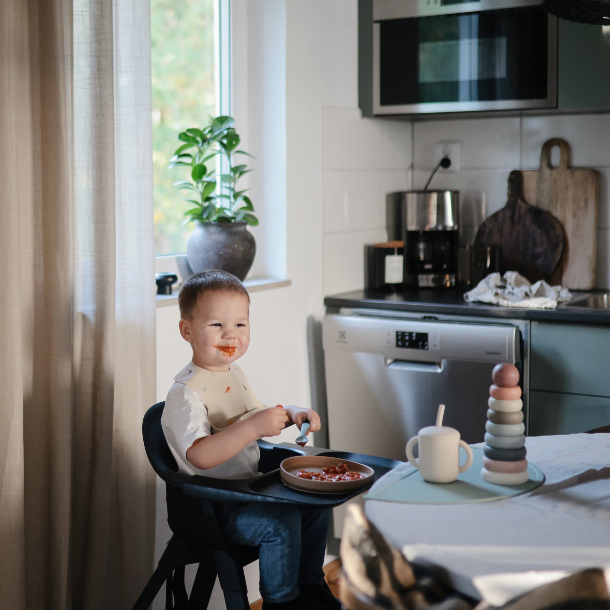 Smiling toddler with mushie Stacking Rings Toy in a cozy Danish Hygge kitchen.
