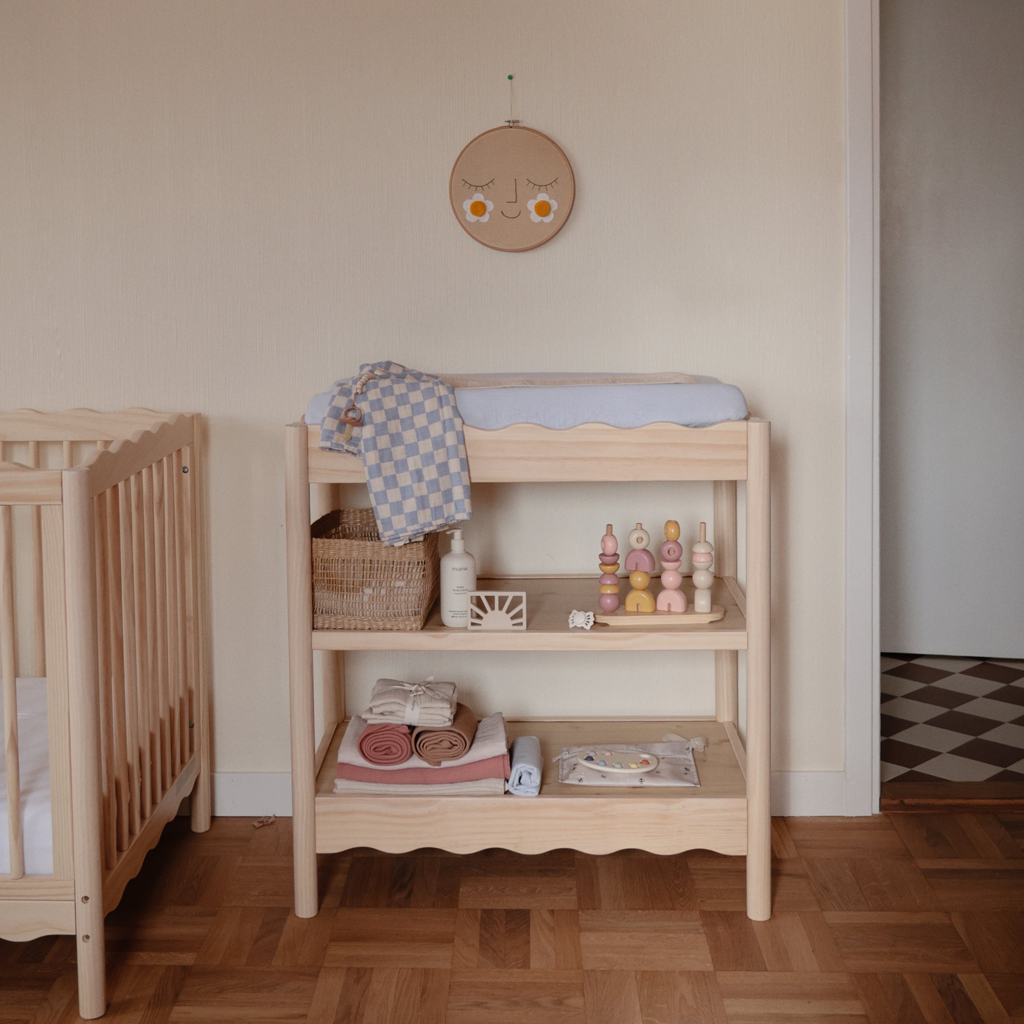 A wooden changing table with folded towels, baby care items, toys, a mushie Sun Teether, and a checkered cloth stands next to a crib in a cozy nursery with wooden flooring and a smiling face wall decoration above.