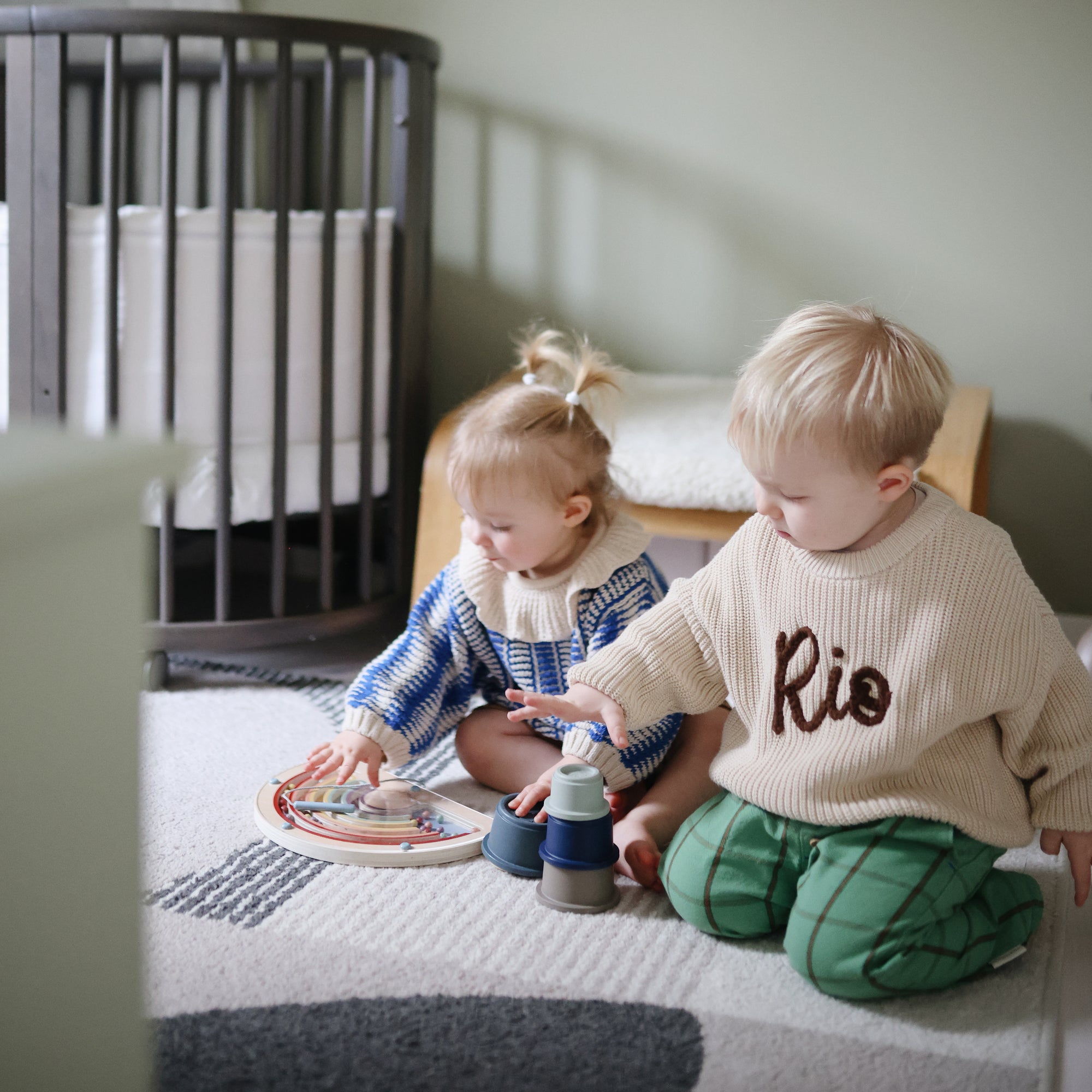 Two young children play with mushie’s Stacking Cups Toy on a rug in a cozy nursery room.