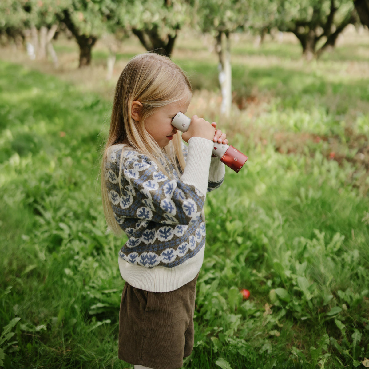 A child plays in a sweater with mushie Kaleidoscopes in a grassy orchard.
