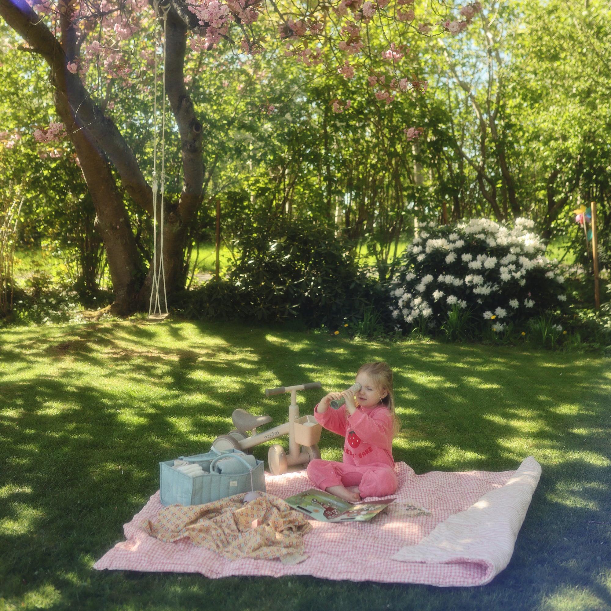 A young girl sits wrapped in a mushie Organic Cotton Muslin Swaddle Blanket on a picnic blanket.