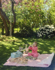 A young girl sits wrapped in a mushie Organic Cotton Muslin Swaddle Blanket on a picnic blanket.