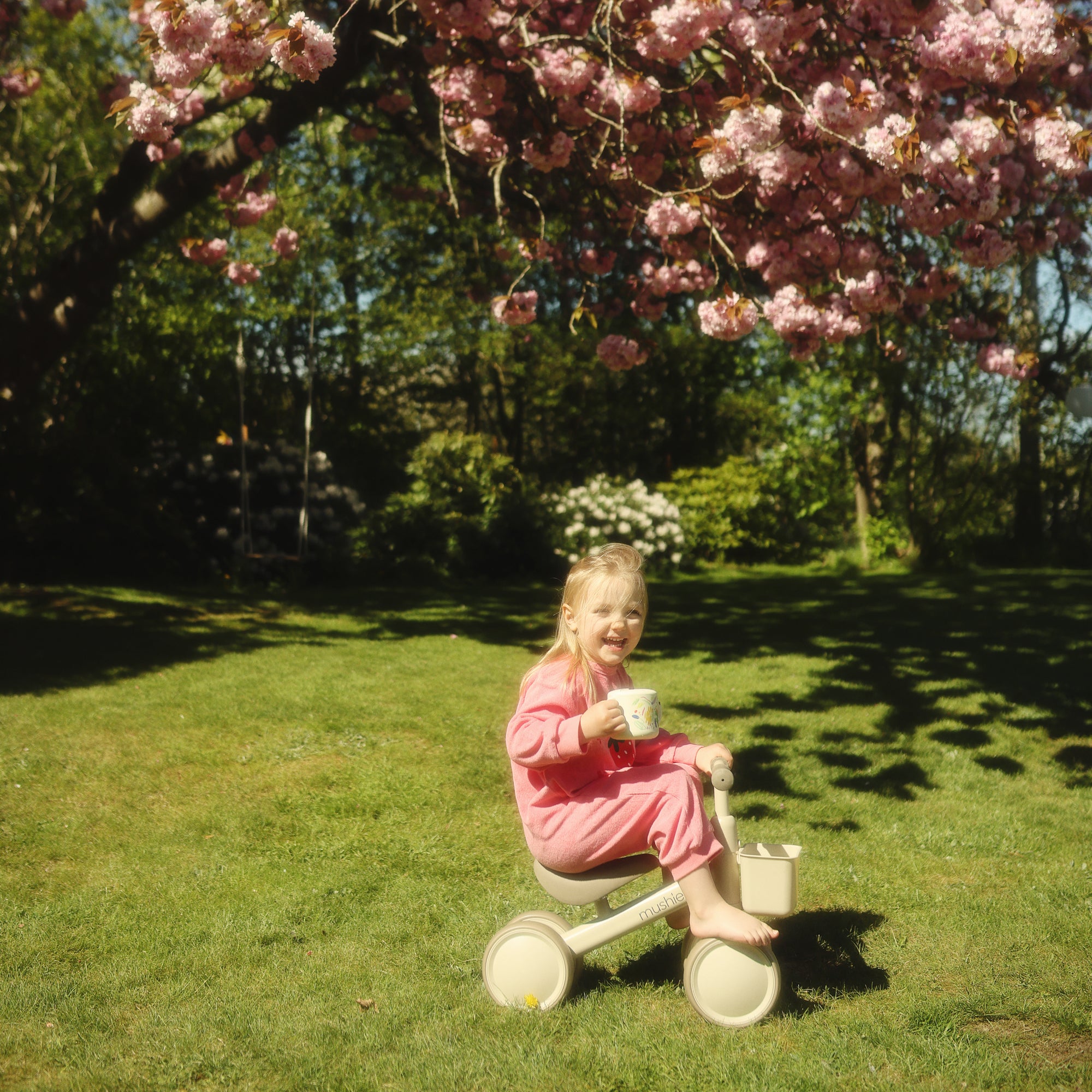 Smiling child in pink pajamas on a mushie Mini Toddler Bike under pink cherry blossoms.
