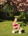 Smiling child in pink pajamas on a mushie Mini Toddler Bike under pink cherry blossoms.