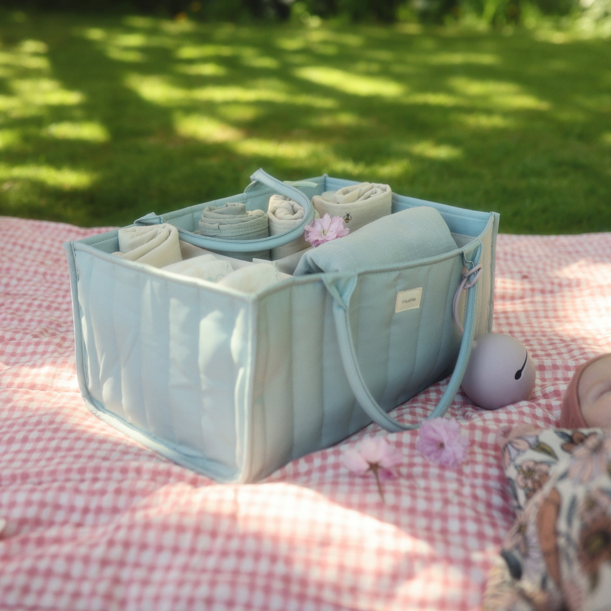 Mushie Diaper Caddy with baby items on a picnic blanket outdoors in sunlight.