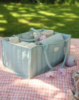 Mushie Diaper Caddy with baby items on a picnic blanket outdoors in sunlight.
