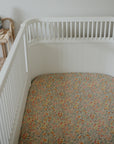 White crib with mushie Extra Soft Muslin Crib Sheet, next to a wicker shelf with pastel rings.