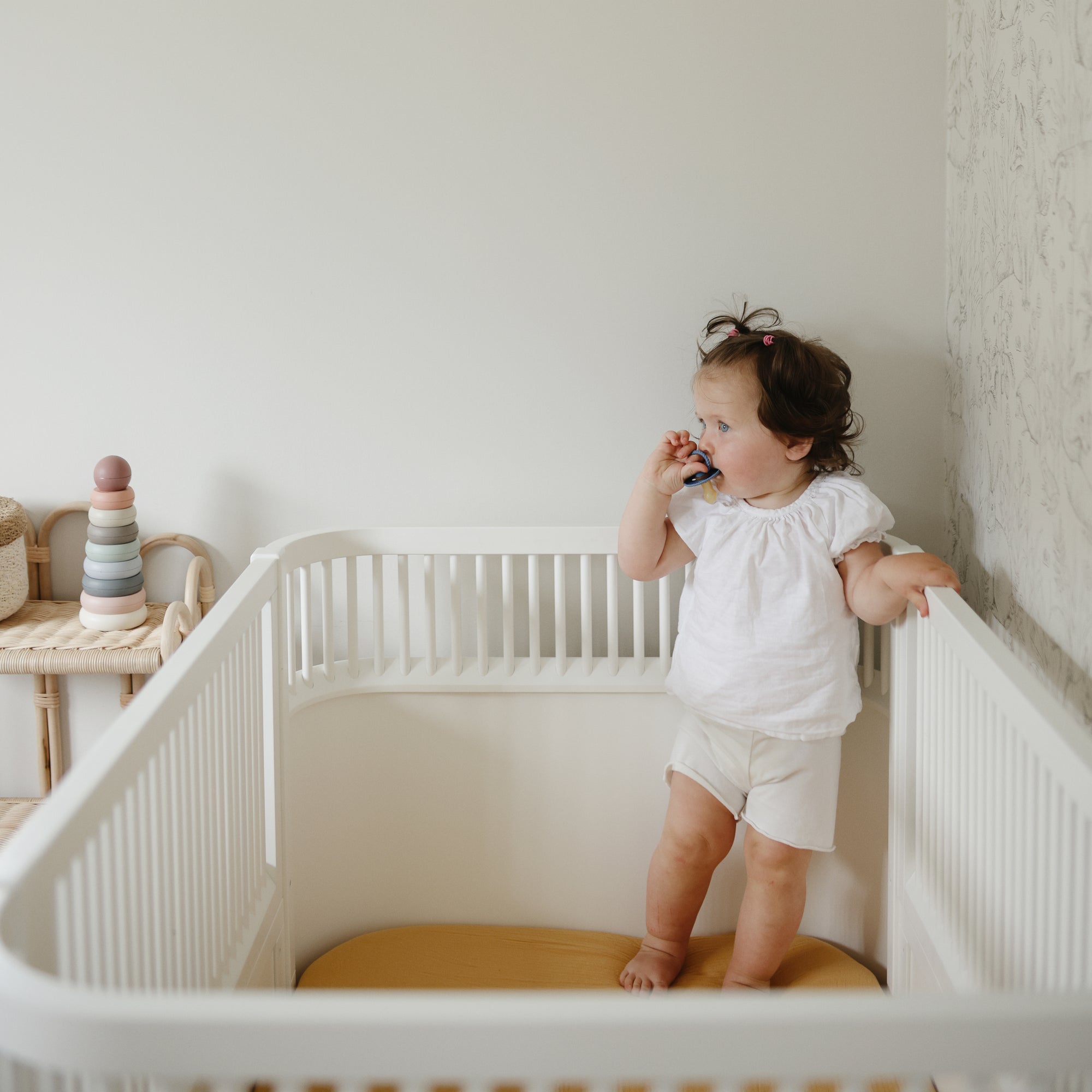 Toddler in a mushie crib with Extra Soft Muslin Crib Sheet, holding a toy and looking aside.