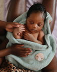 A newborn wrapped in a soft green towel rests on an adult’s lap, with a FRIGG Rope Silicone Pacifier 2-Pack by FRIGG nearby as an adult hand gently touches the baby’s arm—capturing the warmth of true baby essentials.