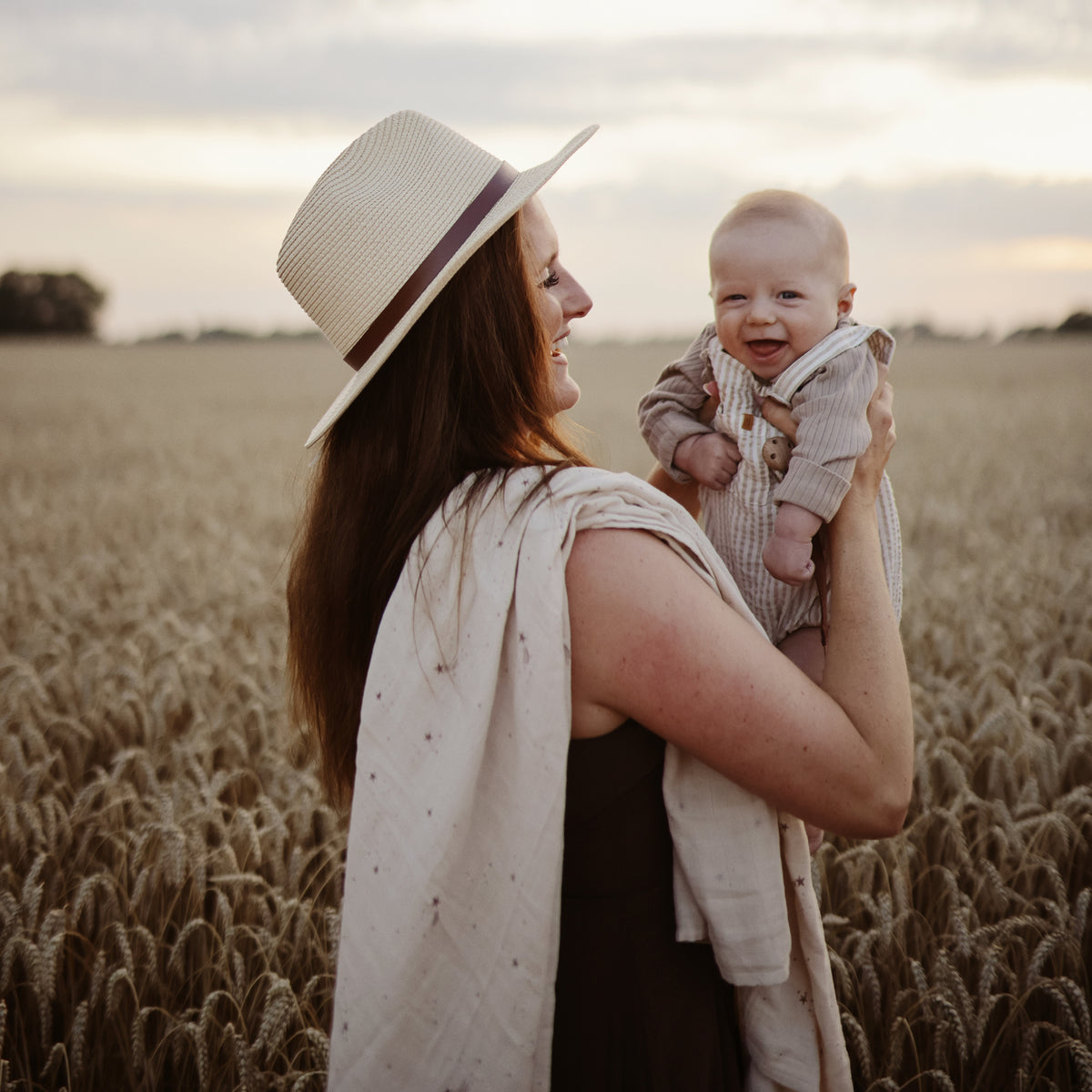 Woman in a hat holds a baby wrapped in a mushie Organic Cotton Muslin Swaddle Blanket at sunset.