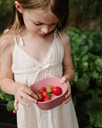 Young girl in a white dress holding mushie Square Dinnerware Bowl in a garden with strawberries.