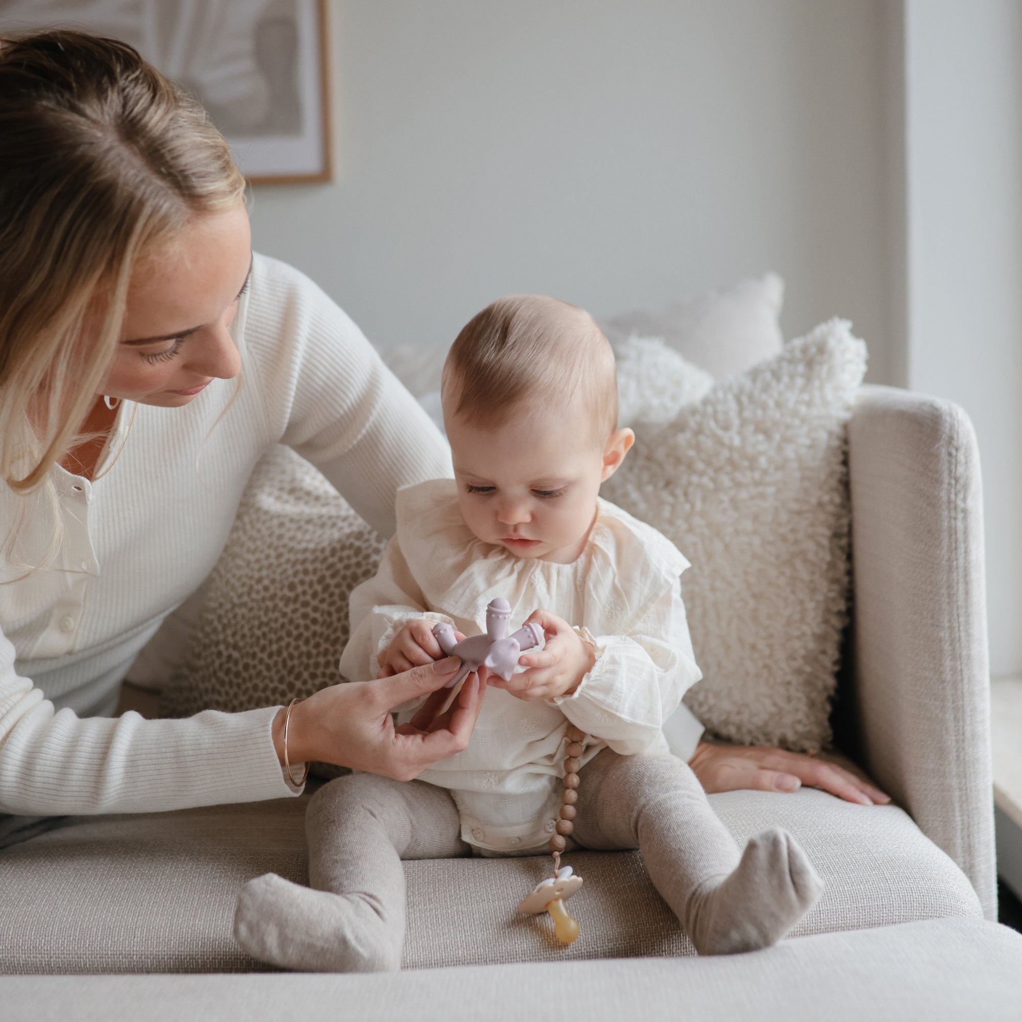 A woman sits on a couch beside a baby, helping the baby hold a purple toy, while a FRIGG Daisy Night Silicone Pacifier from FRIGG rests nearby. The baby, dressed in light-colored clothes, looks down at the toy as the woman gently supports their hands.