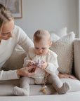 A woman sits on a couch beside a baby, helping the baby hold a purple toy, while a FRIGG Daisy Night Silicone Pacifier from FRIGG rests nearby. The baby, dressed in light-colored clothes, looks down at the toy as the woman gently supports their hands.