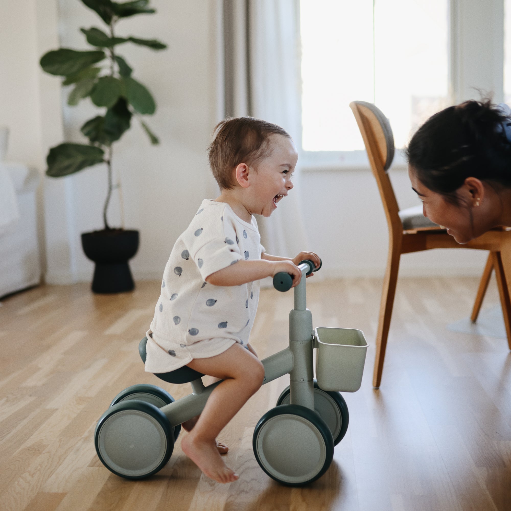A smiling toddler rides a mushie Mini Toddler Bike, laughing at an adult indoors.