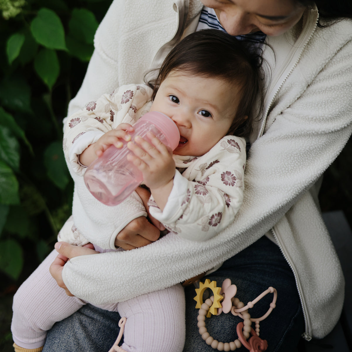 Baby with mushie Soft Spout Sippy Cup on lap, pink, silicone, toys beside.