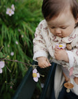 Baby with FRIGG Daisy Pacifier, holding flower by green railing in a garden.