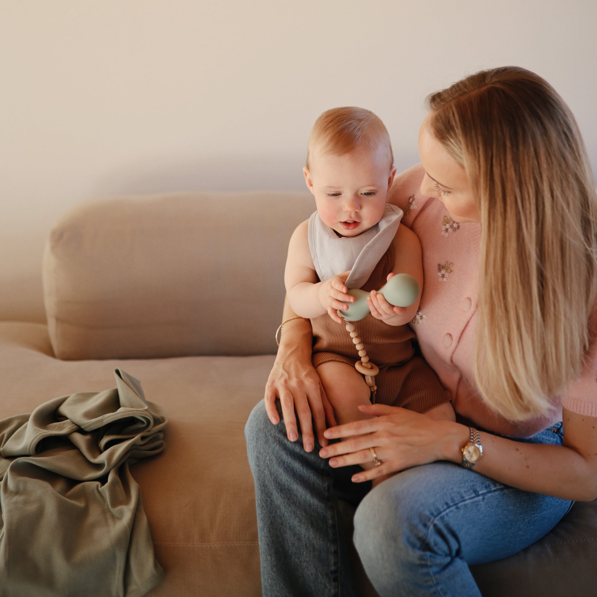 A mother sits on a beige sofa holding her baby, who plays with a sage green silicone teether on a wooden bead chain. Beside them, the Mushie Ribbed Baby Blanket is neatly folded.