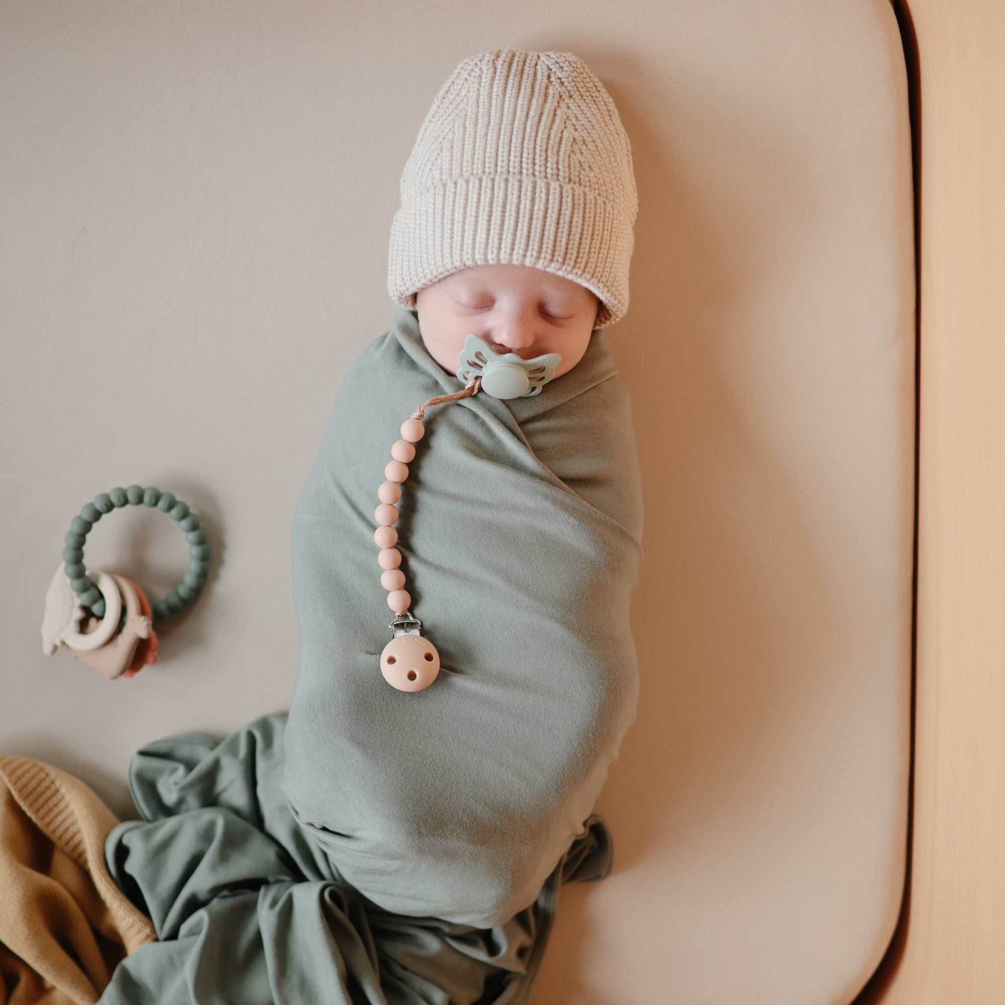 A newborn baby swaddled in a sage green blanket lies on a beige surface, wearing a knit hat and using a pacifier attached to a beaded clip. A teething ring is placed nearby.