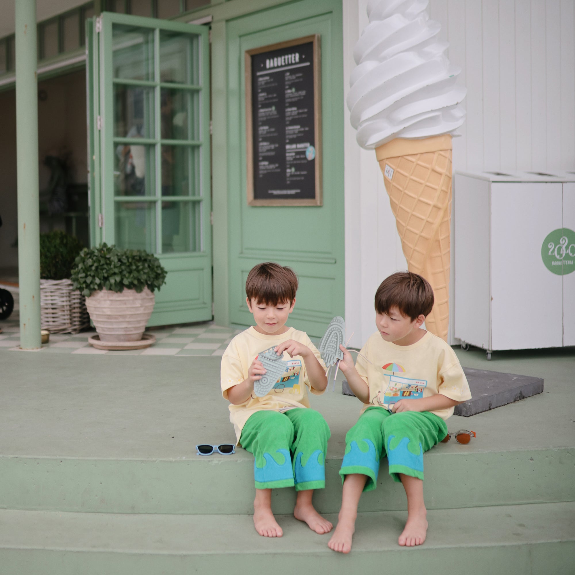 Two kids with mushie Silicone Sensory Toy fidget beside a shop's giant ice cream cone statue.