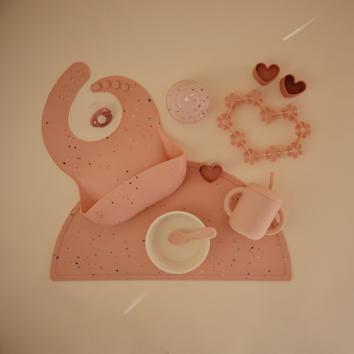 Pink mushie Silicone Baby Bib set with a placemat, bowl, spoon, cup, and toys.