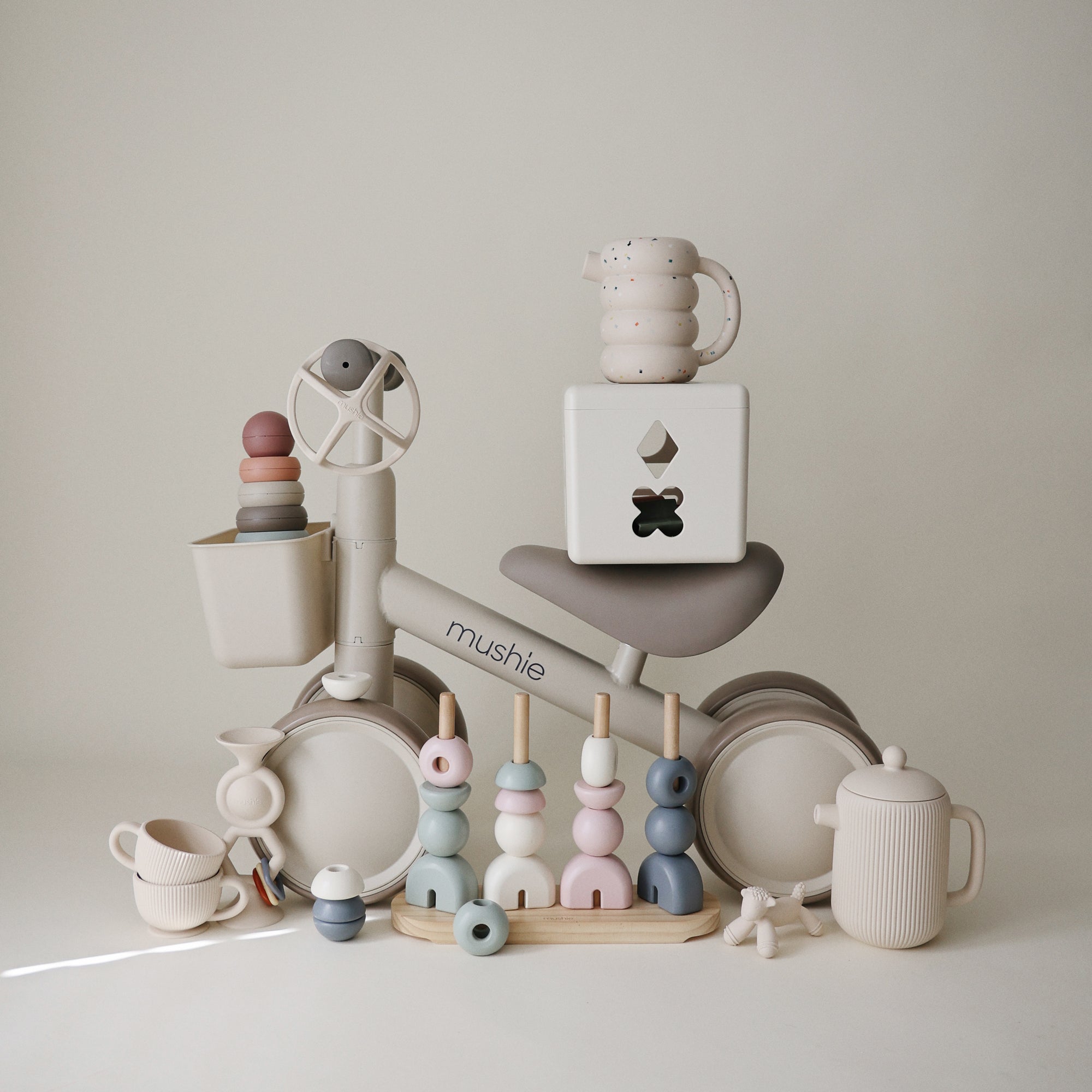 Mushie Mini Toddler Bike with pastel stacking toys, cups, and teapots displayed around it.