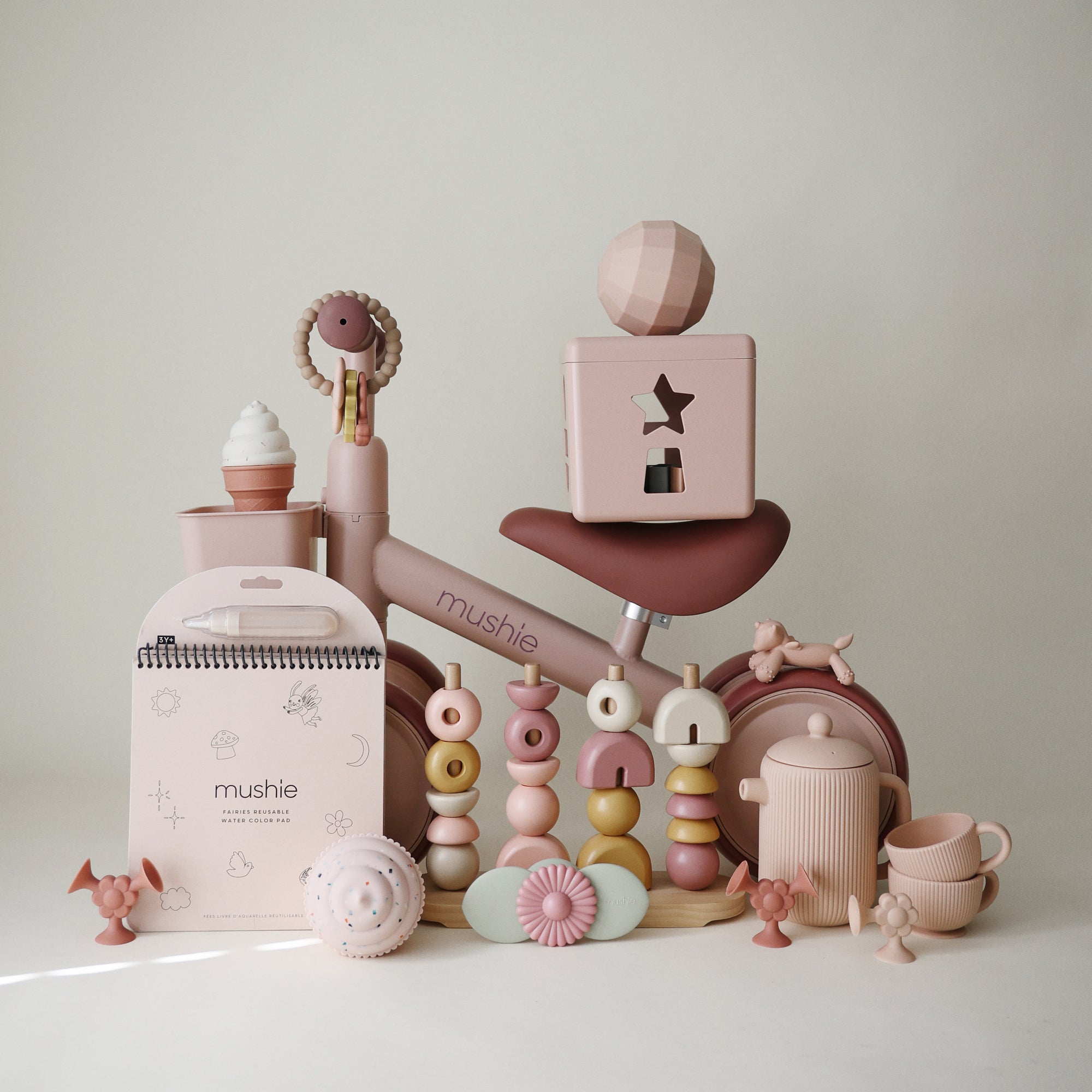 Pastel pink and beige baby toys surround the mushie Mini Toddler Bike.