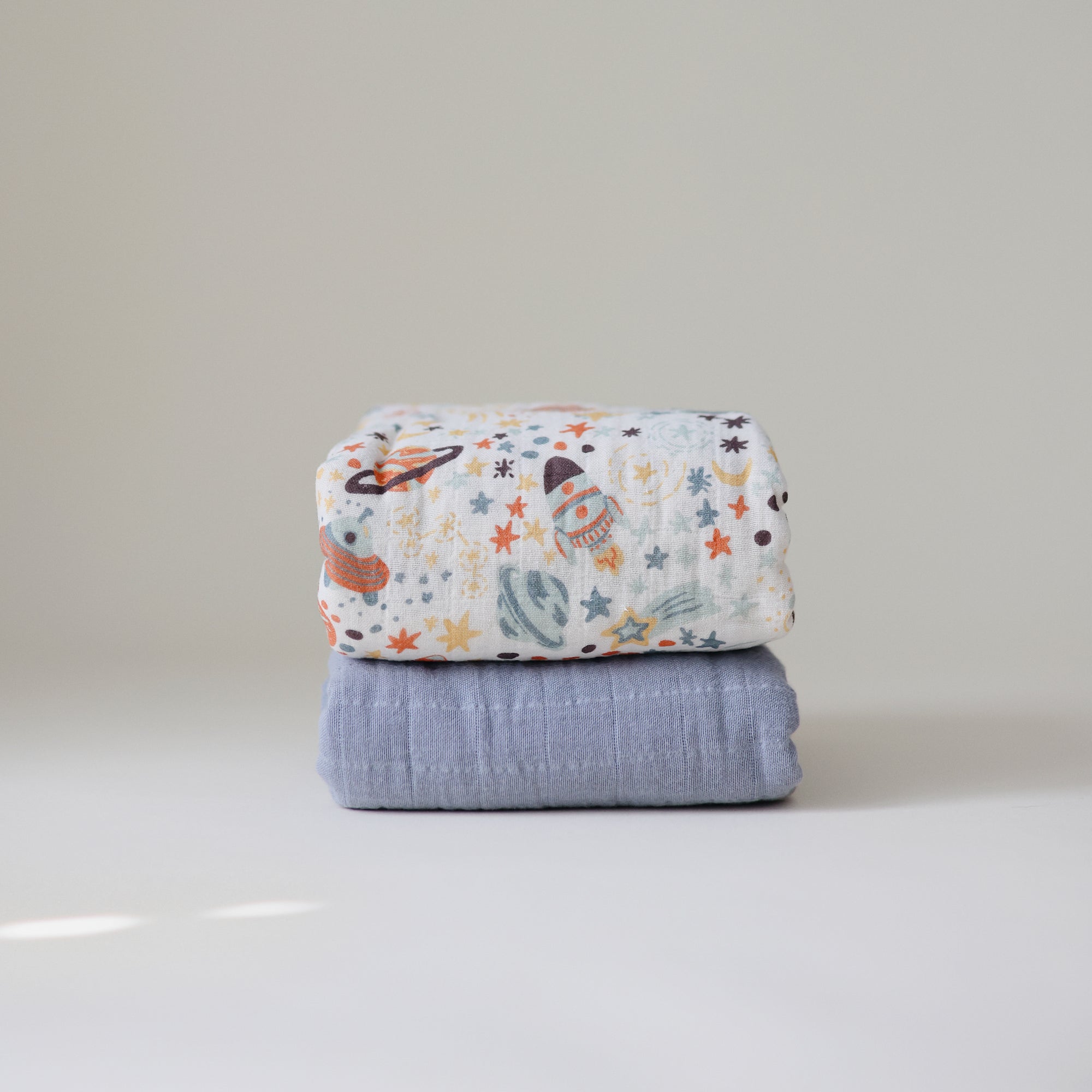 A mushie Organic Cotton Muslin Swaddle Blanket with a space-themed planets and stars print is stacked on a plain light blue baby blanket, both neatly folded against a simple, light background.