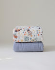 A mushie Organic Cotton Muslin Swaddle Blanket with a space-themed planets and stars print is stacked on a plain light blue baby blanket, both neatly folded against a simple, light background.