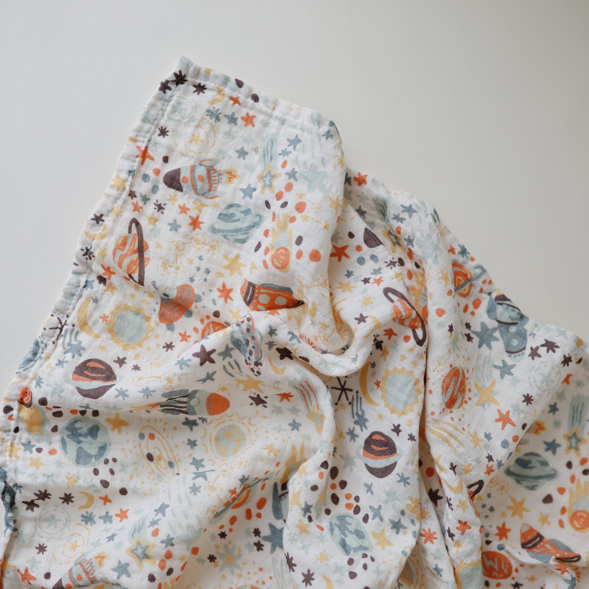 The mushie Organic Cotton Muslin Swaddle Blanket features soft, white organic cotton with colorful prints of planets, stars, and rockets in orange, blue, and brown—perfect for your baby’s comfort.