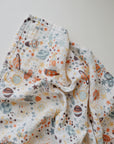 The mushie Organic Cotton Muslin Swaddle Blanket features soft, white organic cotton with colorful prints of planets, stars, and rockets in orange, blue, and brown—perfect for your baby’s comfort.