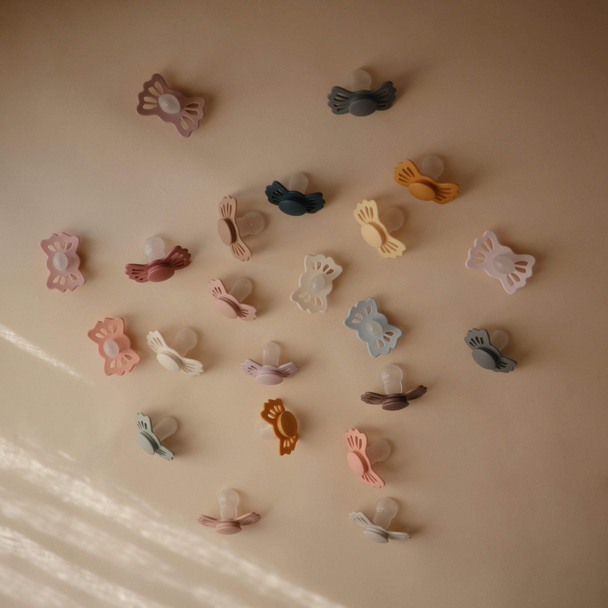 FRIGG Butterfly Anatomical Silicone Pacifier 2-Pack (6-18 Months) by FRIGG features pastel, butterfly-shaped silicone pacifiers in pink, blue, yellow, green, and brown on a beige surface with diagonal light.