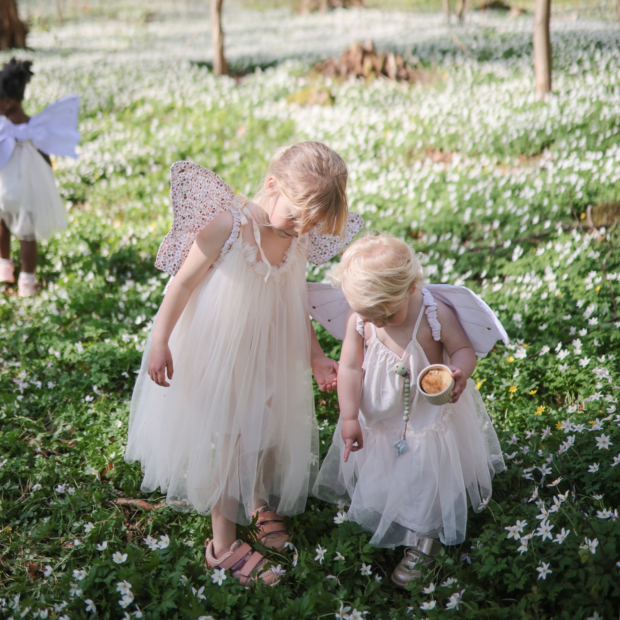 Two young children in white fairy dresses and wings stand among white flowers, one pointing to the ground as they radiate comfort and calm—just like FRIGG Butterfly Anatomical Silicone Pacifier 2-Pack (6-18 Months) from FRIGG. Another child in wings appears behind them.