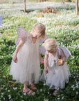 Two young children in white fairy dresses and wings stand among white flowers, one pointing to the ground as they radiate comfort and calm—just like FRIGG Butterfly Anatomical Silicone Pacifier 2-Pack (6-18 Months) from FRIGG. Another child in wings appears behind them.