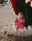 A girl smiles on a playground, holding the mushie Silicone Sensory Toy in her mouth.